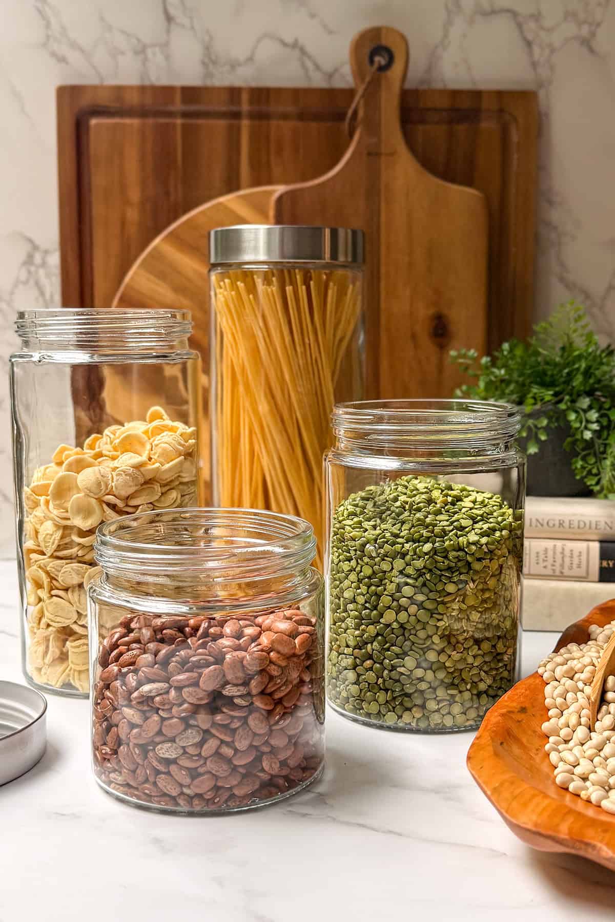 Dried pinto beans, split peas, and pasta in clear glass storage jars with wooden cutting boards in the background.