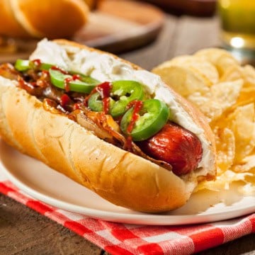 A Seattle-Style Hot Dog topped with cream cheese, saut&eacute;ed onions, jalape&ntilde;o slices, and hot sauce, served on a plate with crispy potato chips.