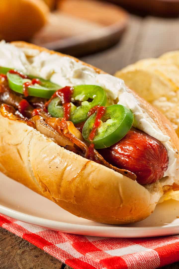 A Seattle-Style Hot Dog topped with cream cheese, sautéed onions, jalapeño slices, and hot sauce, served on a plate with crispy potato chips.