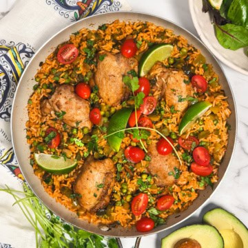 One skillet arroz con pollo with chicken thighs, rice, peas, tomatoes, and lime wedges.