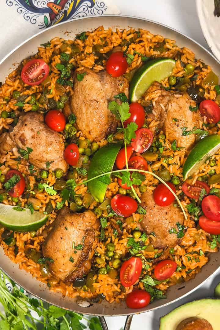One skillet arroz con pollo with chicken thighs, rice, peas, tomatoes, and lime wedges.