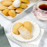 Three madeleines dusted with powdered sugar on a white plate, with a tray of more madeleines and a cup of tea in the background—perfect inspiration for trying your own madeleines recipe at home.