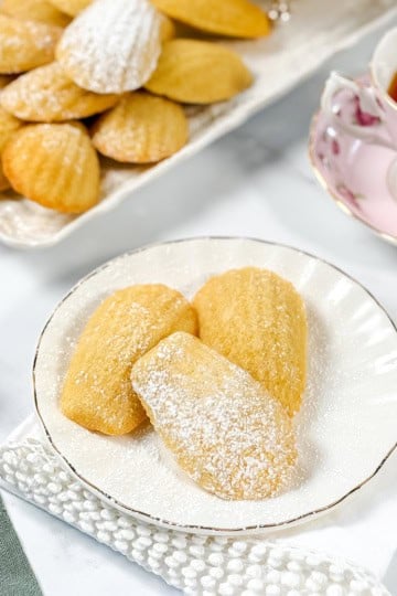 Three madeleines dusted with powdered sugar on a white plate, with a tray of more madeleines and a cup of tea in the background—perfect inspiration for trying your own madeleines recipe at home.