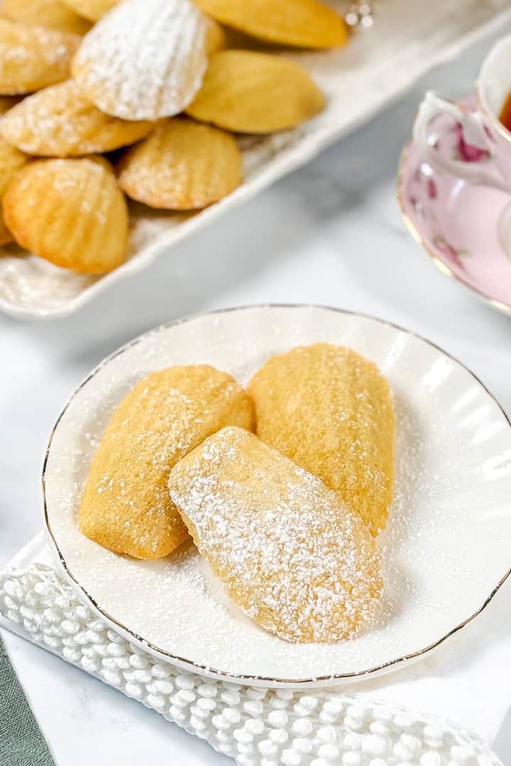 Three madeleines dusted with powdered sugar on a white plate, with a tray of more madeleines and a cup of tea in the background-perfect inspiration for trying your own madeleines recipe at home.