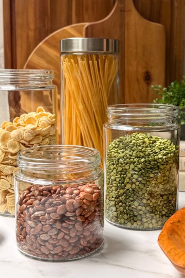 Dried beans, legumes, and pasta in clear glass storage jars with wooden cutting boards in the background.
