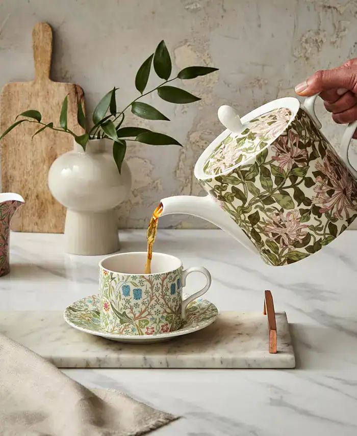 A hand pours tea from a Spode William Morris and Co floral-patterned teapot into a matching teacup on a saucer, set on a marble tray-an intimate moment that speaks the language of love, with a vase, leaf stem, and wooden board in the background.
