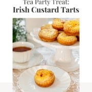 Irish Custard Tarts displayed on a white cake stand and a plate, next to a cup of tea, rest elegantly on a lace tablecloth beneath.