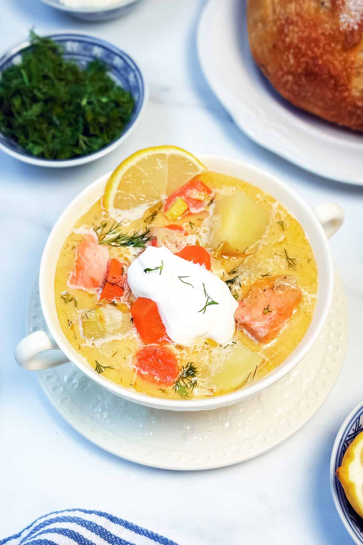 A bowl of Finnish Salmon Soup (Lohikeitto) with chunks of salmon, potatoes, carrots, fresh dill, a lemon slice, and a dollop of sour cream on top, served on a white plate.