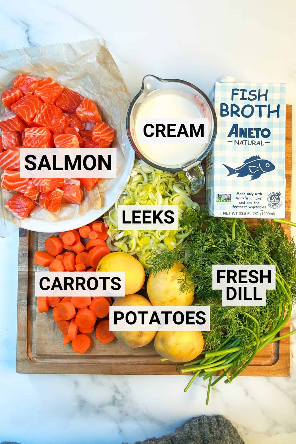 Chopped salmon, carrots, leeks, potatoes, fresh dill, a measuring cup of cream, and a carton of fish broth arranged on a wooden board-classic ingredients for Lohikeitto, traditional Finnish Salmon Soup.
