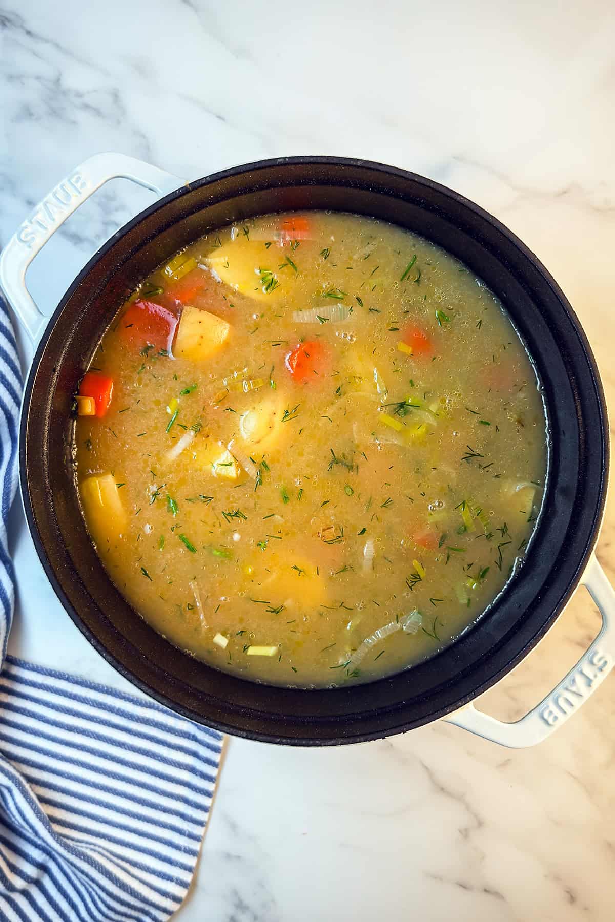 A pot of Lohikeitto, or Finnish Salmon Soup, with visible chunks of potatoes, carrots, salmon, and herbs sits on a marble surface next to a striped cloth.