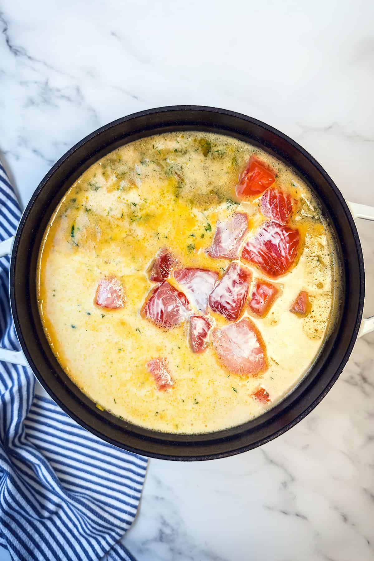 A pot of creamy Finnish Salmon Soup (Lohikeitto) with chopped tomatoes on top, set on a marble surface next to a blue and white striped cloth.