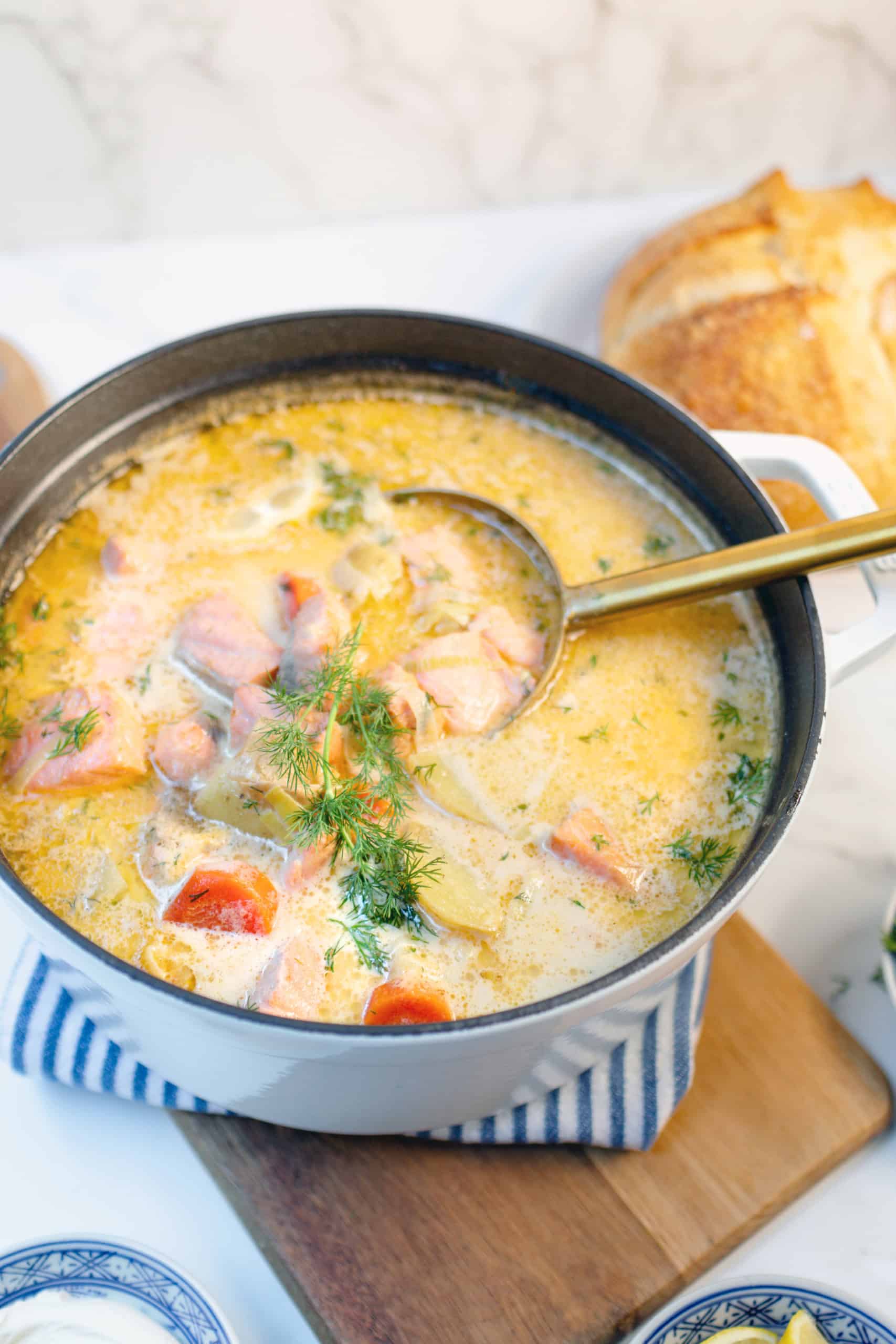 A pot of creamy Finnish Salmon Soup, or Lohikeitto, with chunks of salmon, potatoes, and carrots, garnished with fresh dill, sits on a wooden board with a ladle and bread in the background.