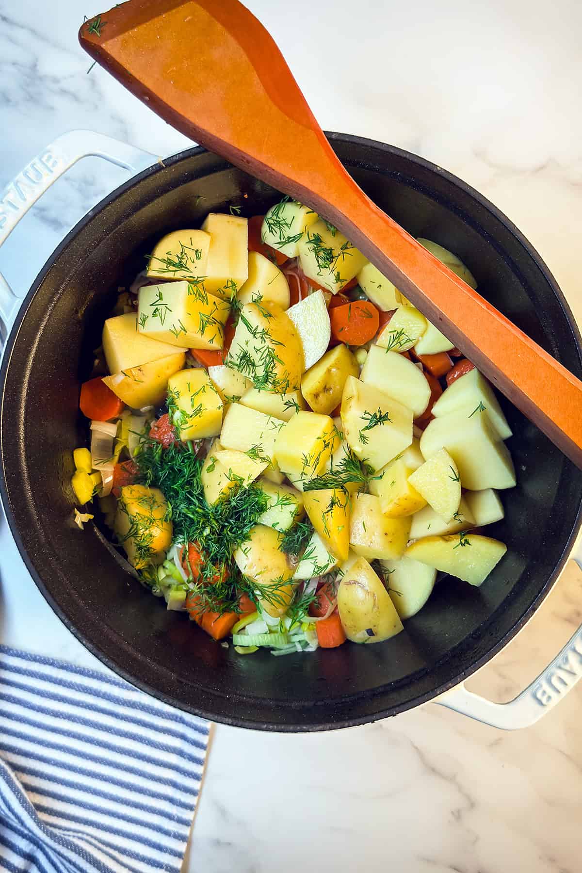 A pot filled with chopped potatoes, carrots, and herbs for Finnish Salmon Soup rests on a marble surface, a wooden spatula balanced on top beside a striped cloth.