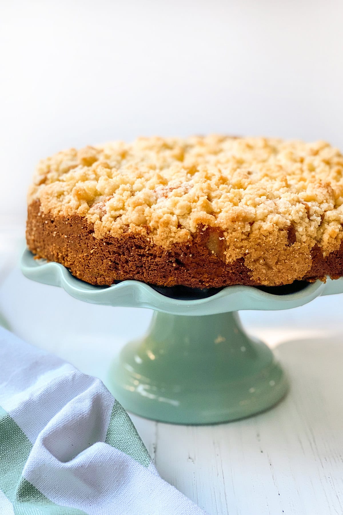 An Irish apple crumble cake sits on a light green pedestal cake stand, its crumb topping inviting, with a striped cloth nearby on a white wooden surface.