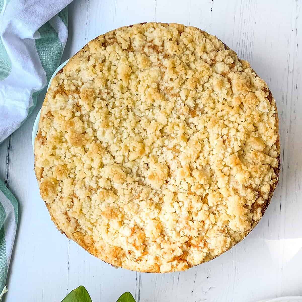 A baked Irish apple crumble cake sits on a white wooden table beside a green plant, two forks, and a white plate.