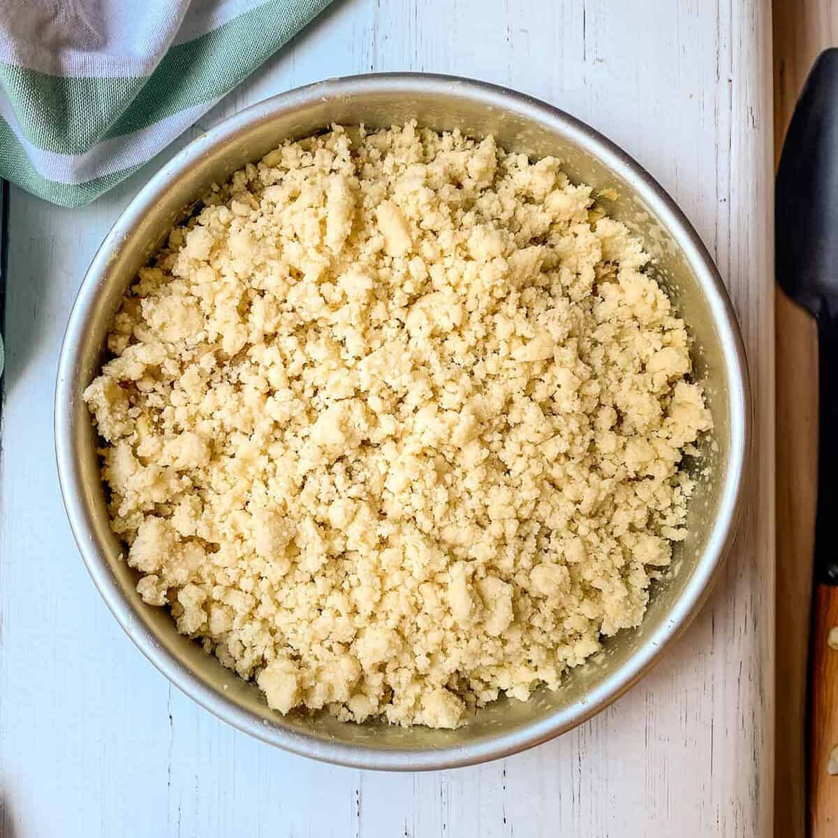 Metal bowl filled with crumble topping for the Irish Apple Crumble Cake sits on a white wooden surface next to a green-striped towel and a spatula.