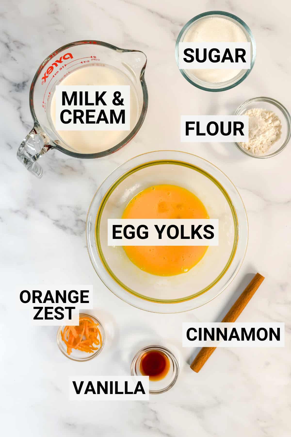 Bowls and containers with labeled ingredients-milk & cream, sugar, flour, egg yolks, orange zest, vanilla, and a cinnamon stick-are arranged on a marble surface, ready to be used in Irish Custard Tarts.