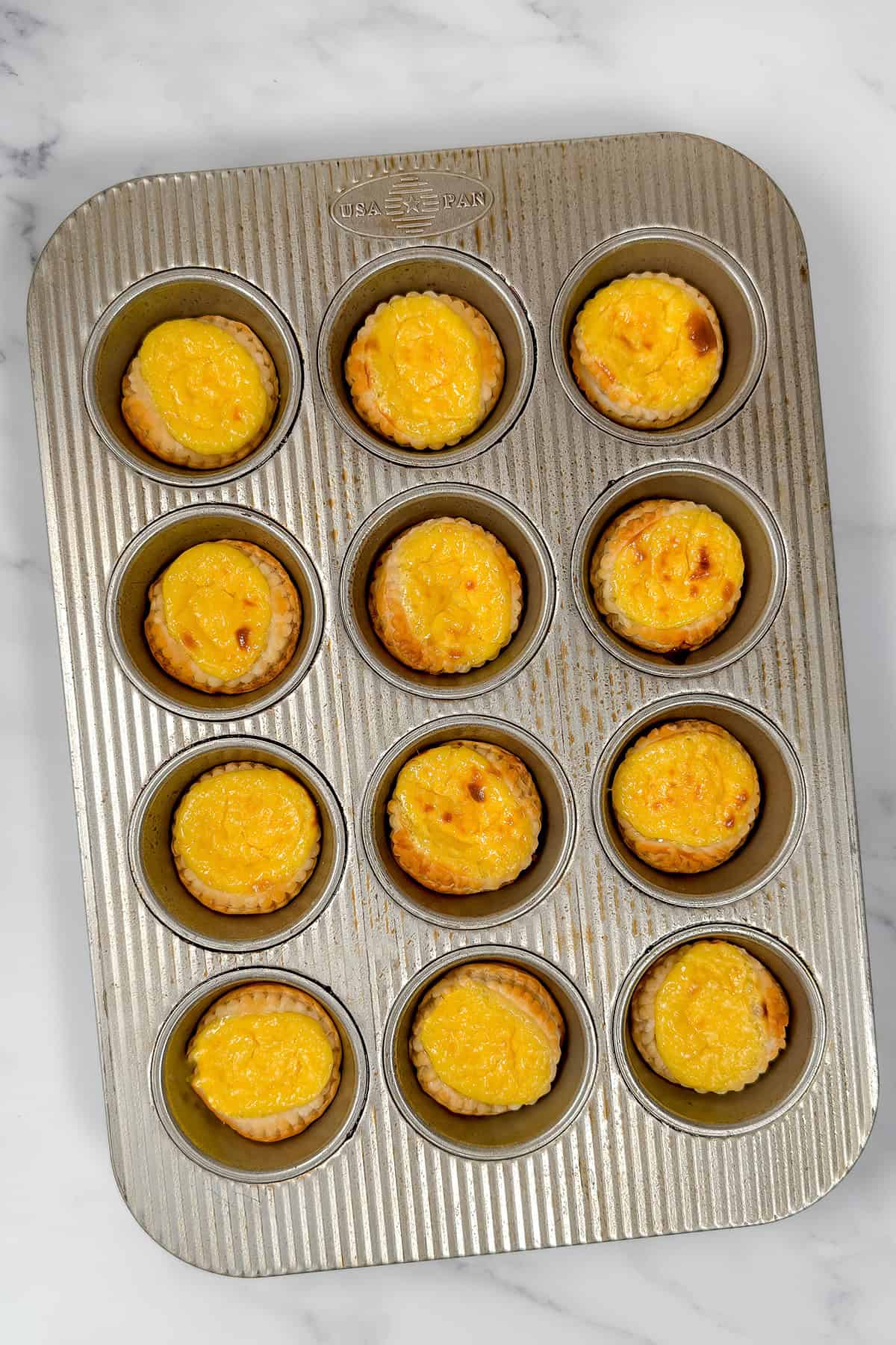 A muffin tin holding twelve golden baked Irish Custard Tarts on a marble surface, perfect for anyone craving classic Irish desserts.