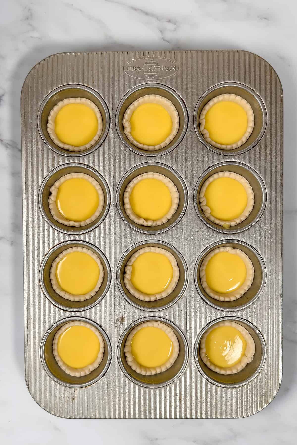 A metal muffin tray holds twelve unbaked Irish Custard Tarts with yellow filling on a white marble surface.