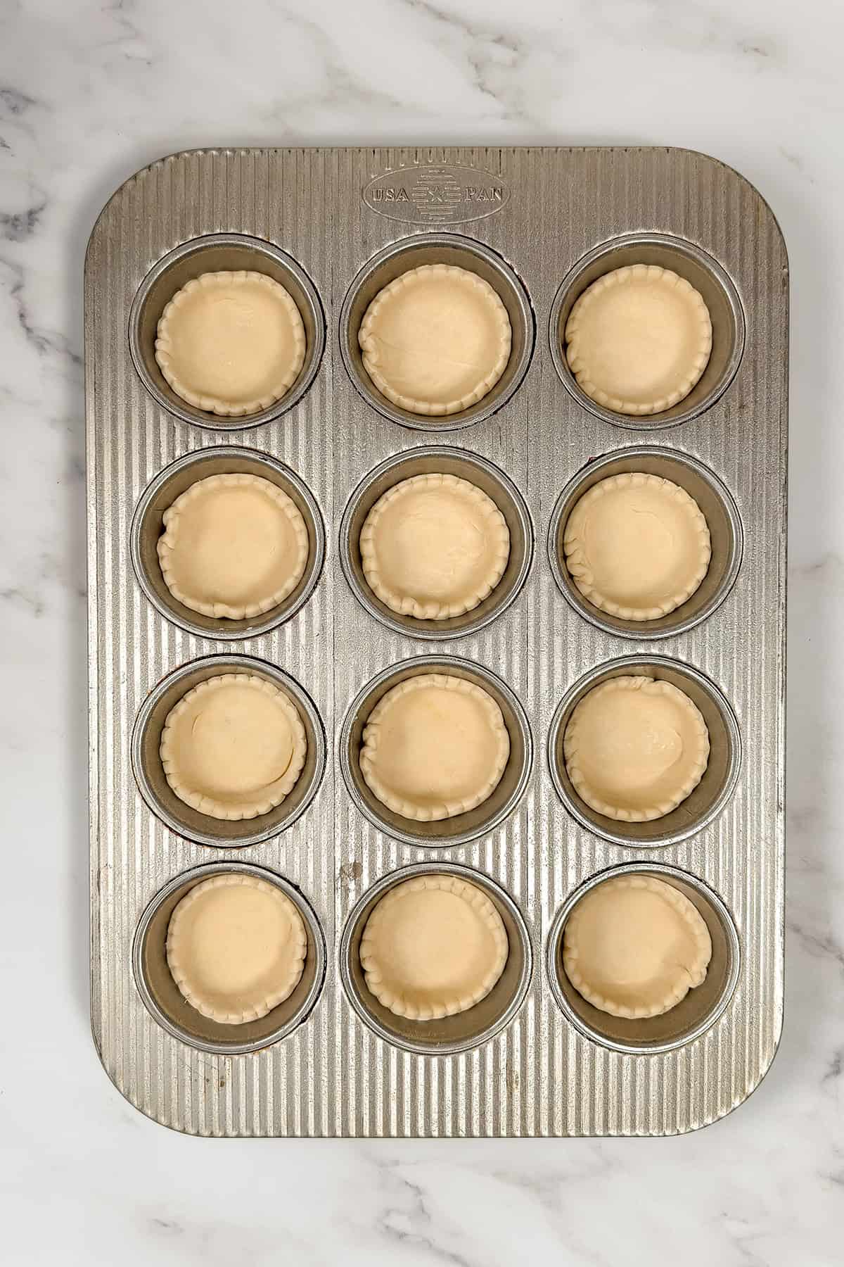 A muffin tin with twelve unbaked Irish custard tart pastry crusts arranged in each cup, placed on a white marble surface.