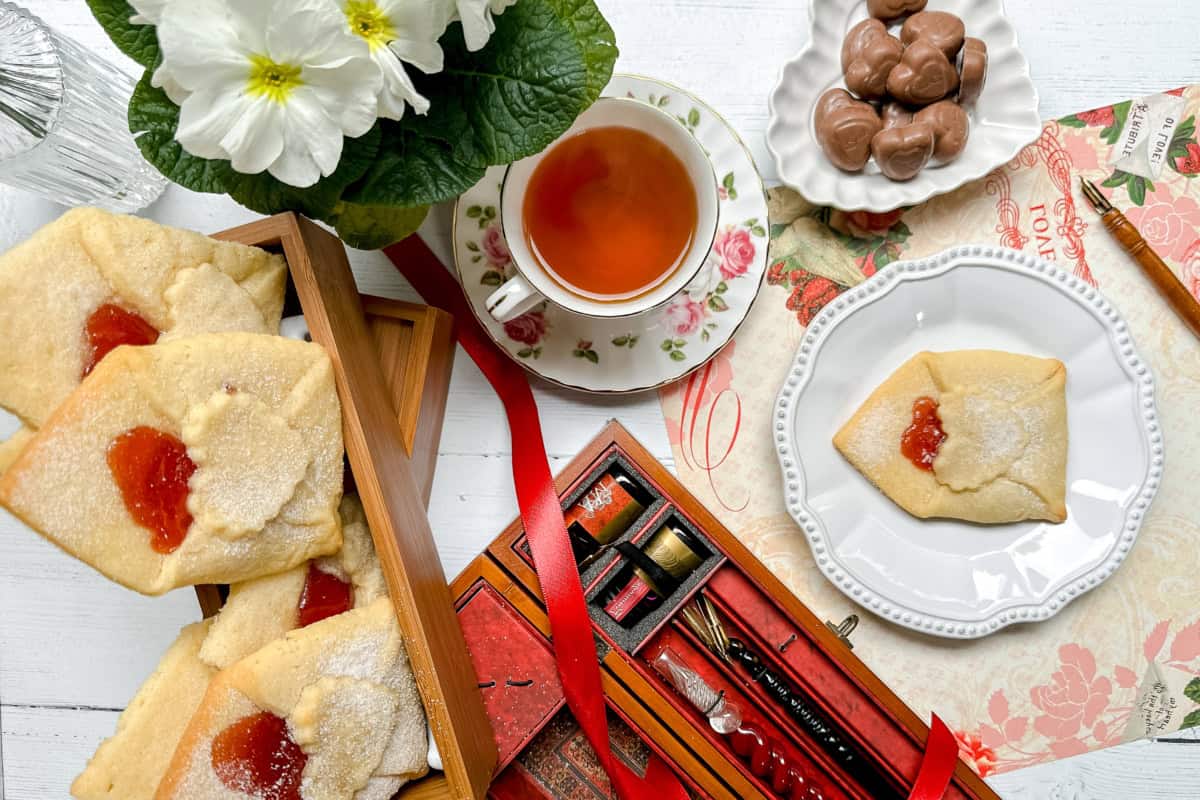 A tea set with a floral teacup, a plate of jam-filled love letter cookies, chocolate candies, a potted plant, and a red box of calligraphy pens sits ready on the white table-perfect for relaxing before game day excitement begins.
