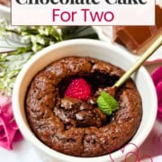 A mini chocolate cake recipe served in a white ramekin, topped with a raspberry and mint, with a gold spoon beside. Chocolate squares and flowers sit in the background. Text overlay reads "Mini Lava Style Chocolate Cake For Two.