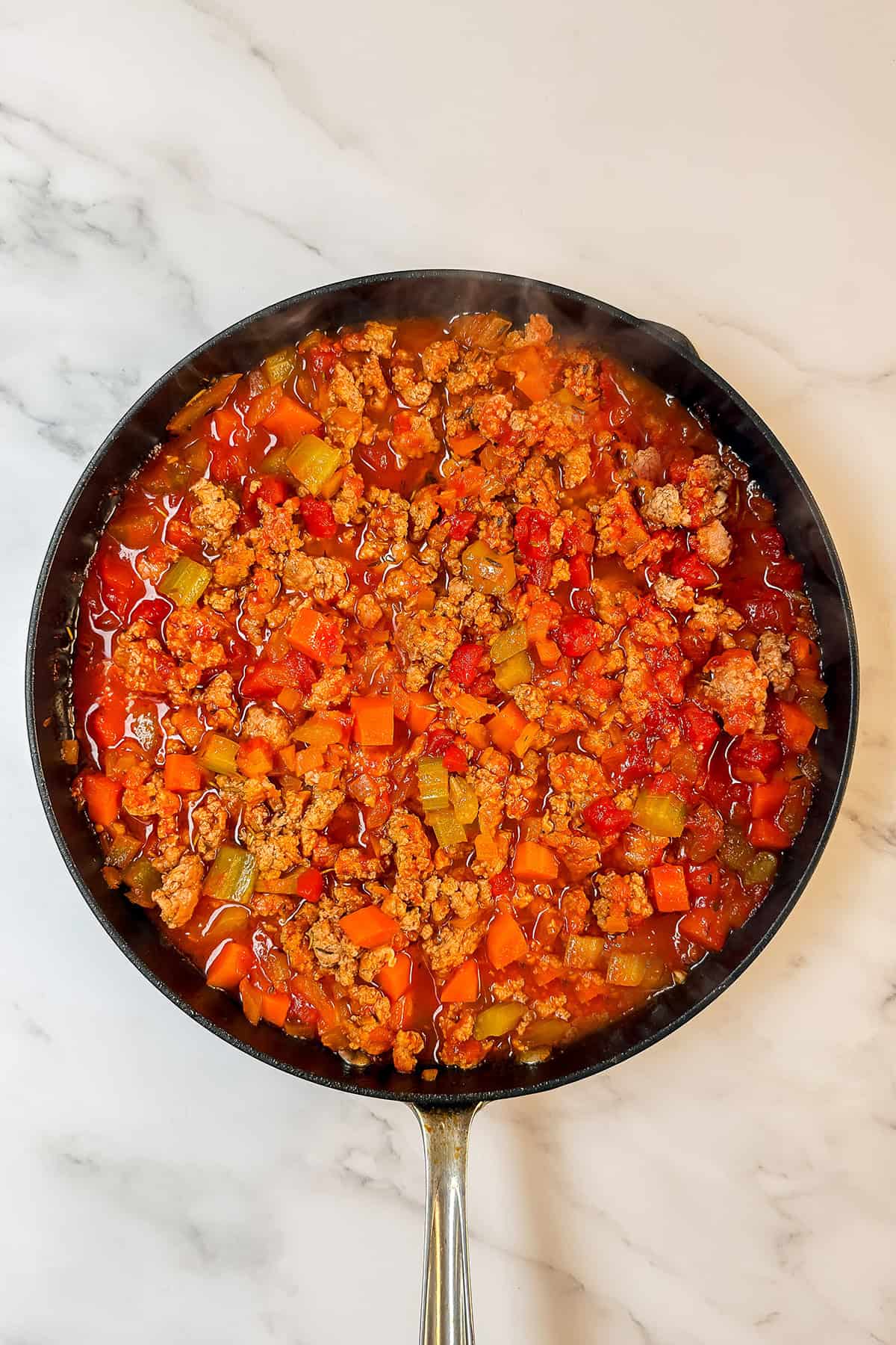 A Quick Skillet filled with ground meat, diced tomatoes, carrots, celery, and peppers simmers in a red sauce on a marble countertop-perfect for easy 30 Minute Recipes.