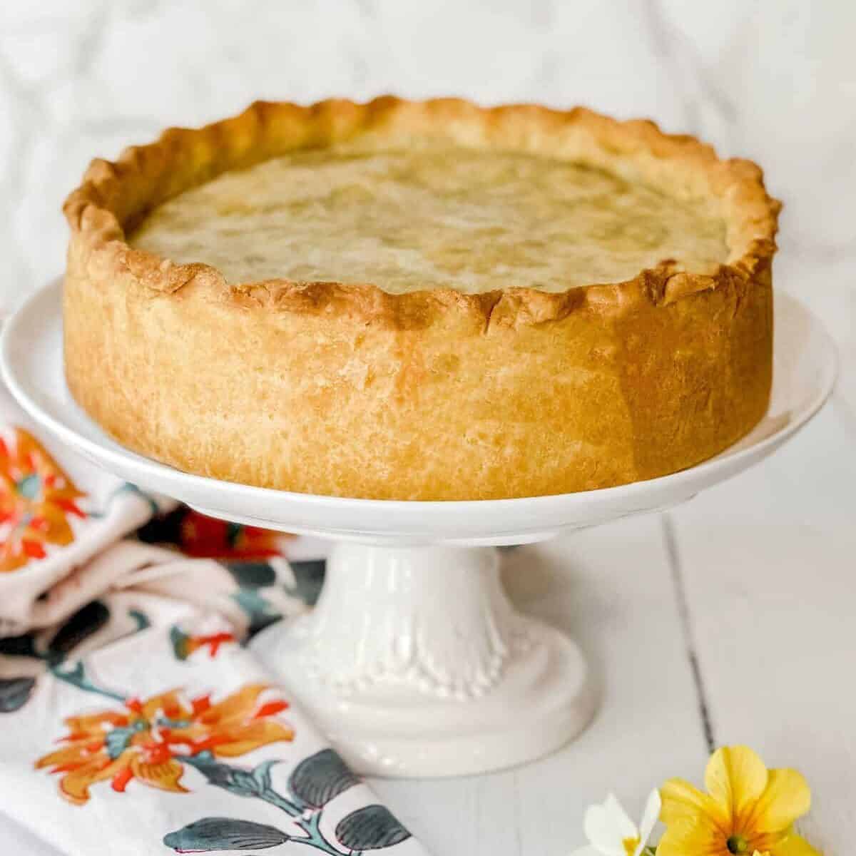 A deep-dish pie, reminiscent of a classic pizza rustica, boasts a golden-brown crust on a white cake stand. A floral-patterned cloth and yellow and white flowers decorate the white wooden surface, set against a light marble-textured background.