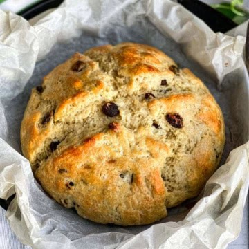 A round loaf of Irish soda bread sits on crumpled parchment paper in a baking dish, freshly baked with a golden-brown crust.