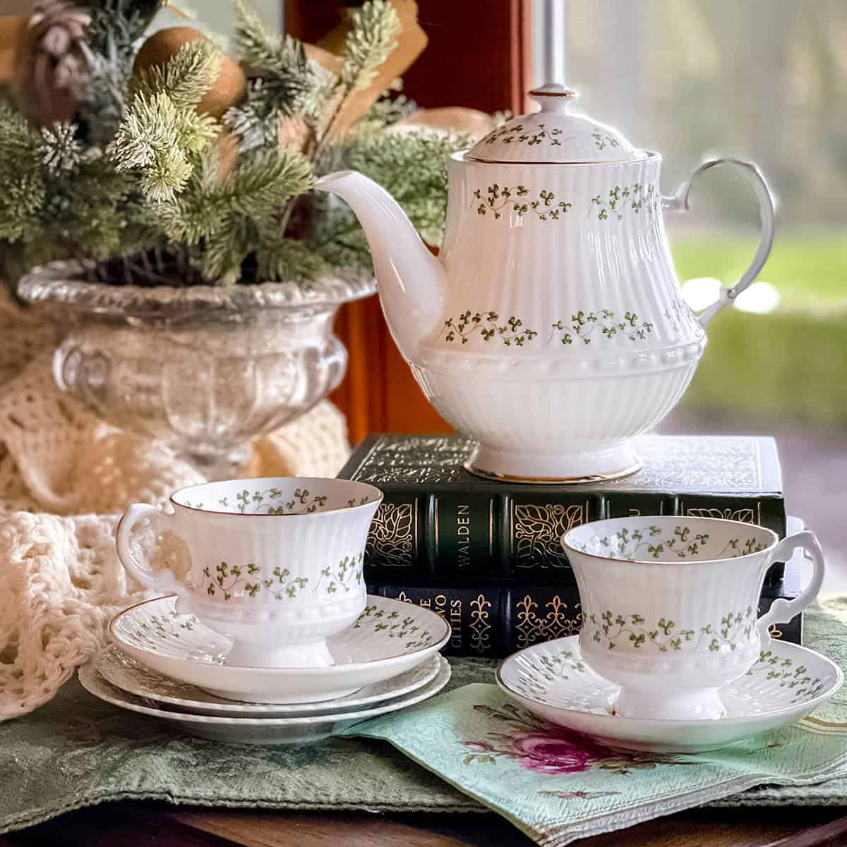 A white teapot and two matching teacups with green clover patterns are arranged on stacked books near a window, setting the scene for afternoon tea by month, with greenery and a crocheted blanket in the background.