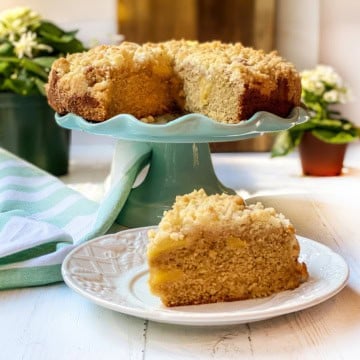 An Irish apple crumble cake sits on a turquoise cake stand, its crumb topping perfectly golden, with a slice served on a white plate in the foreground.