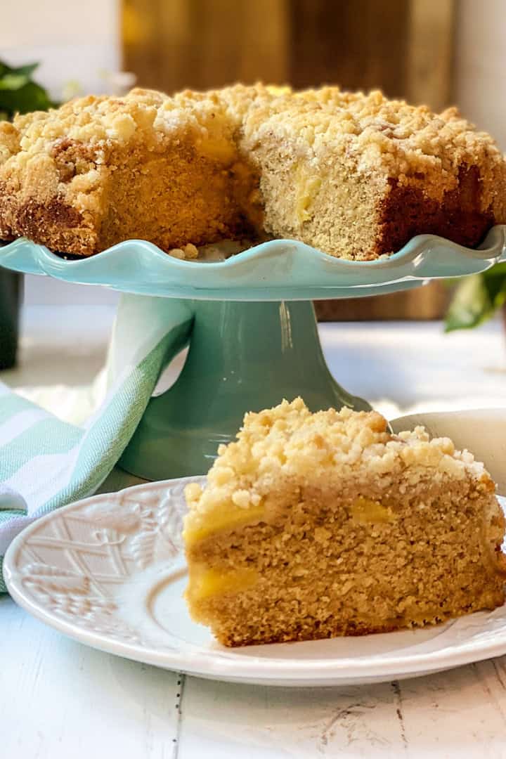 An Irish apple crumble cake sits on a turquoise cake stand, its crumb topping perfectly golden, with a slice served on a white plate in the foreground.