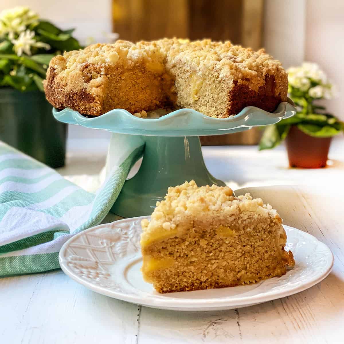 An Irish apple crumble cake sits on a turquoise cake stand, its crumb topping perfectly golden, with a slice served on a white plate in the foreground.