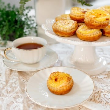 A plate with an Irish Custard Tart sits in front of a teacup filled with tea, while more egg tarts are arranged on a cake stand in the background.
