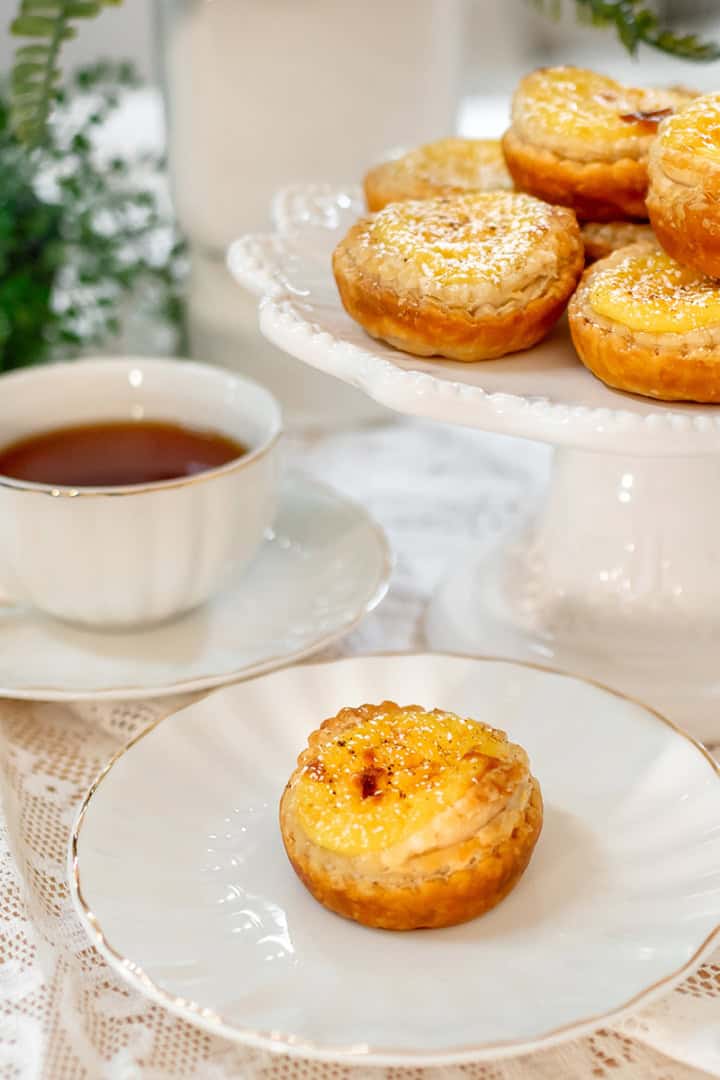 A plate with an Irish Custard Tart sits in front of a teacup filled with tea, while more egg tarts are arranged on a cake stand in the background.