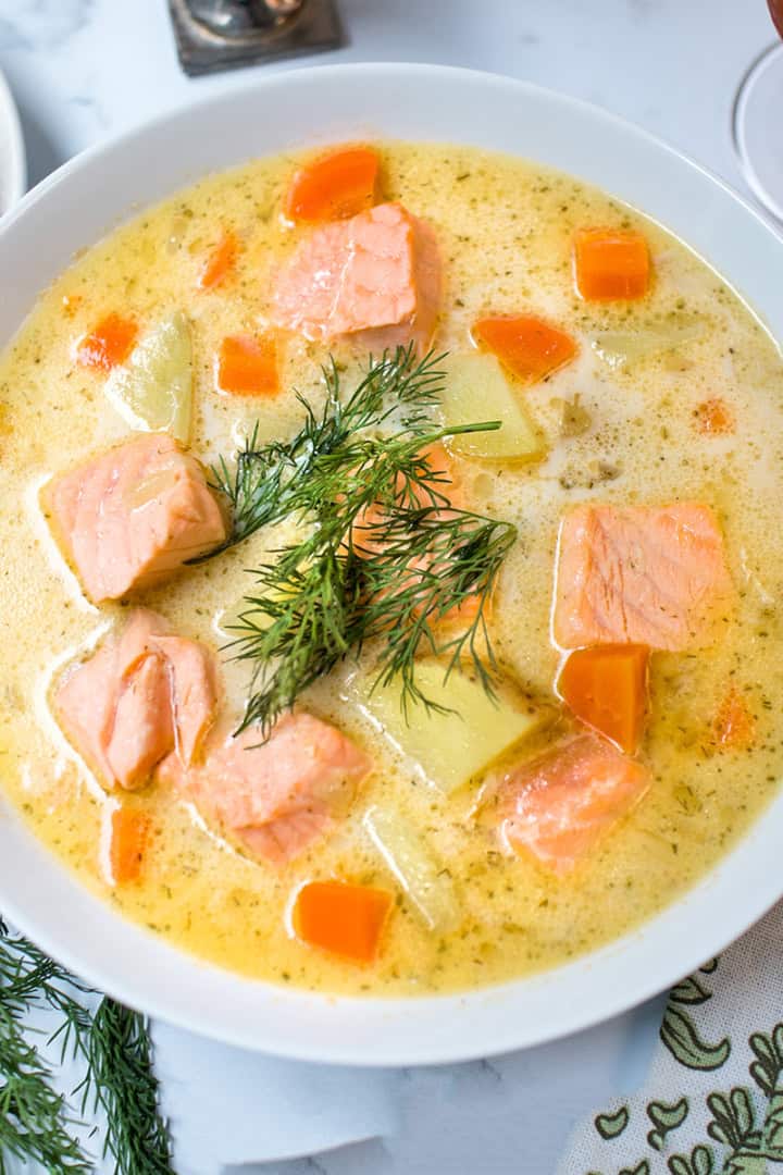 A bowl of creamy Finnish Salmon Soup with chunks of salmon, carrots, and potatoes, garnished with fresh dill, sits on a table beside a slice of bread.