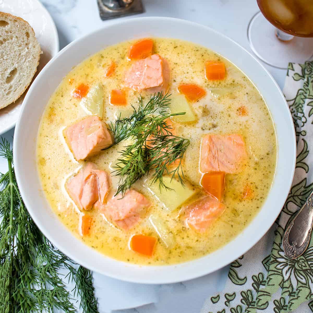 A bowl of creamy Finnish Salmon Soup with chunks of salmon, carrots, and potatoes, garnished with fresh dill, sits on a table beside a slice of bread.