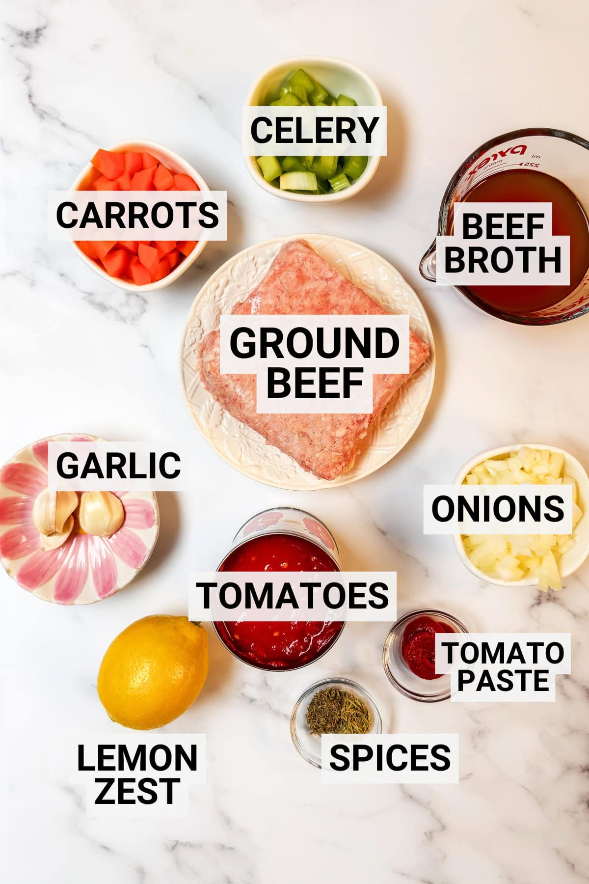 Labeled ingredients for a 30-Minute Osso Buco recipe arranged on a white surface, including ground beef, carrots, celery, beef broth, garlic, onions, tomatoes, tomato paste, lemon zest, and spices.