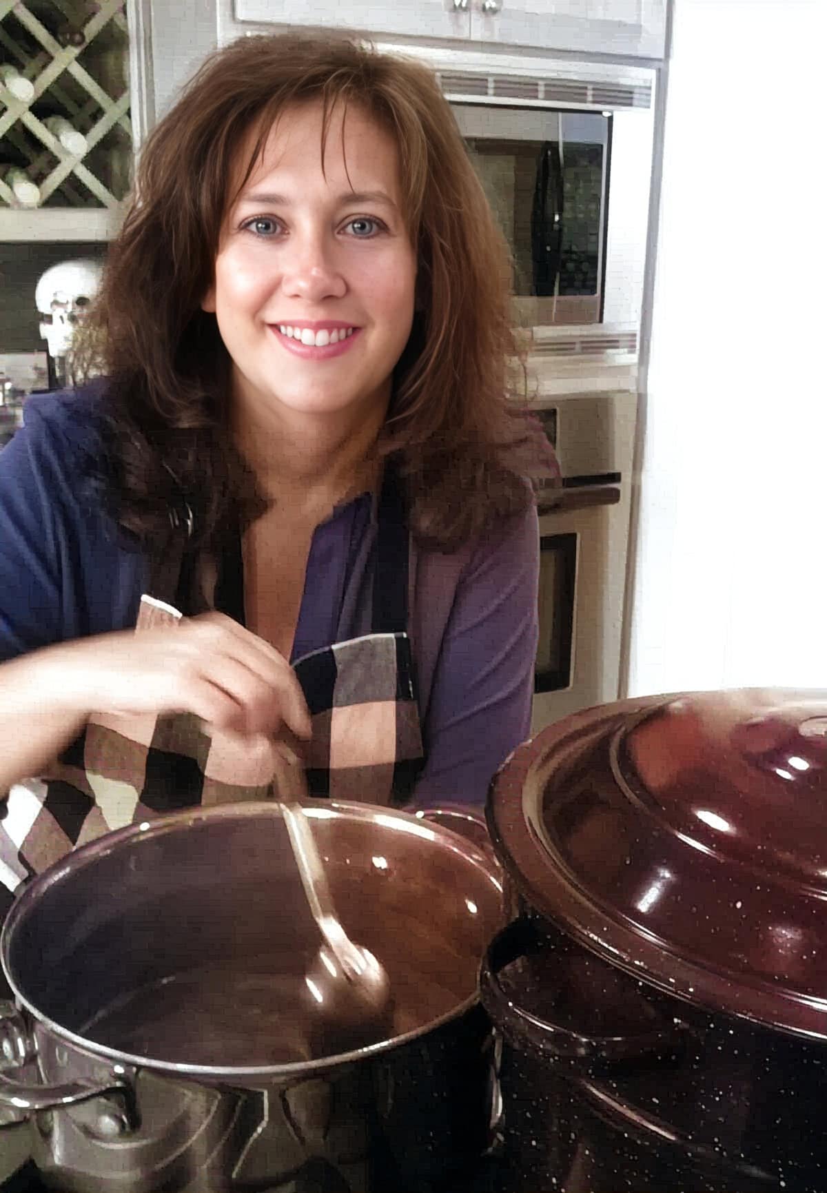 A woman in an apron stirs a pot on a kitchen counter, with a large covered pot next to her and appliances in the background-capturing the warmth of About 31 Daily home cooking.