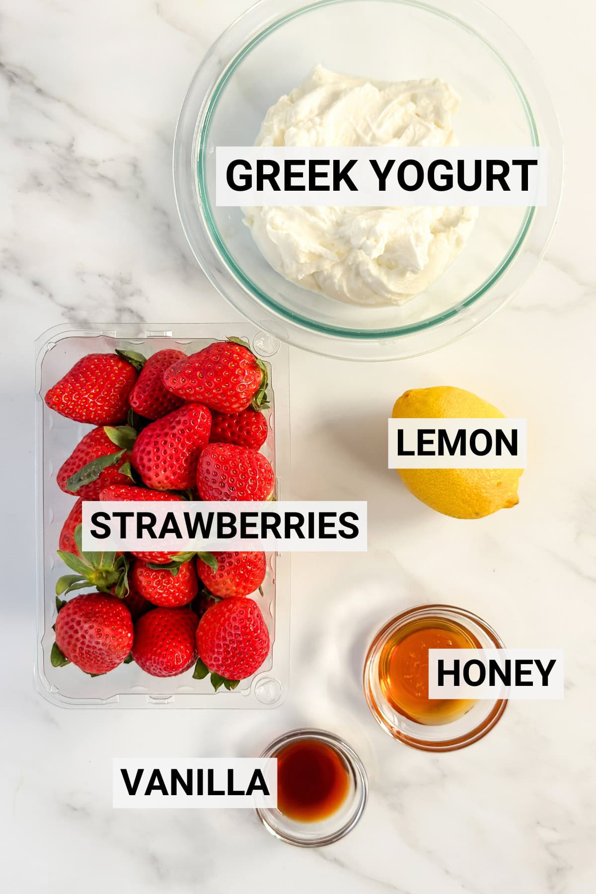 A bowl of Greek yogurt, a lemon, a container of strawberries, a small bowl of honey-sweetened syrup, and a small bowl of vanilla rest on a marble surface.