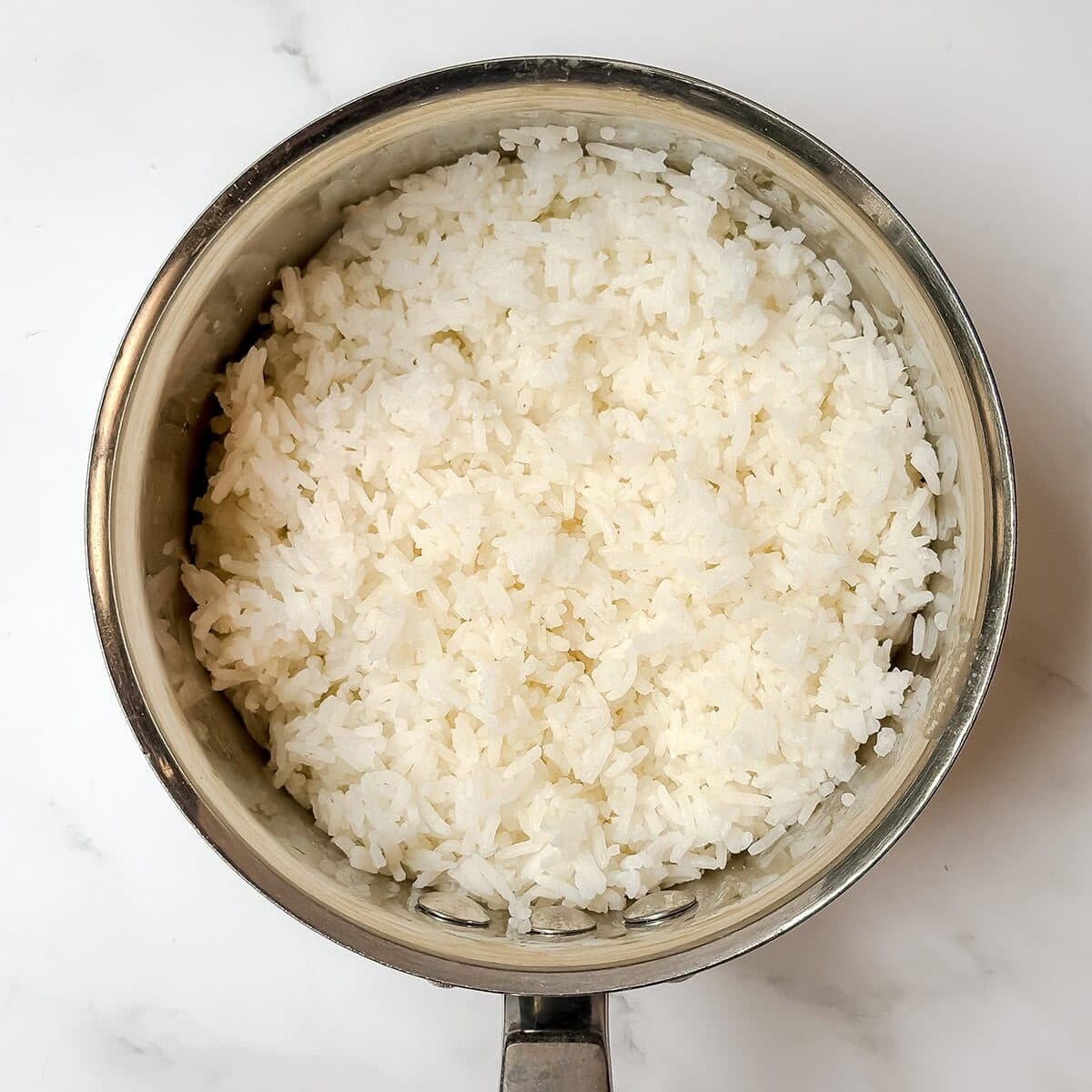 A stainless steel saucepan filled with cooked white rice, perfect for Shawarma Bowls or other quick chicken recipes, placed on a white marble surface.