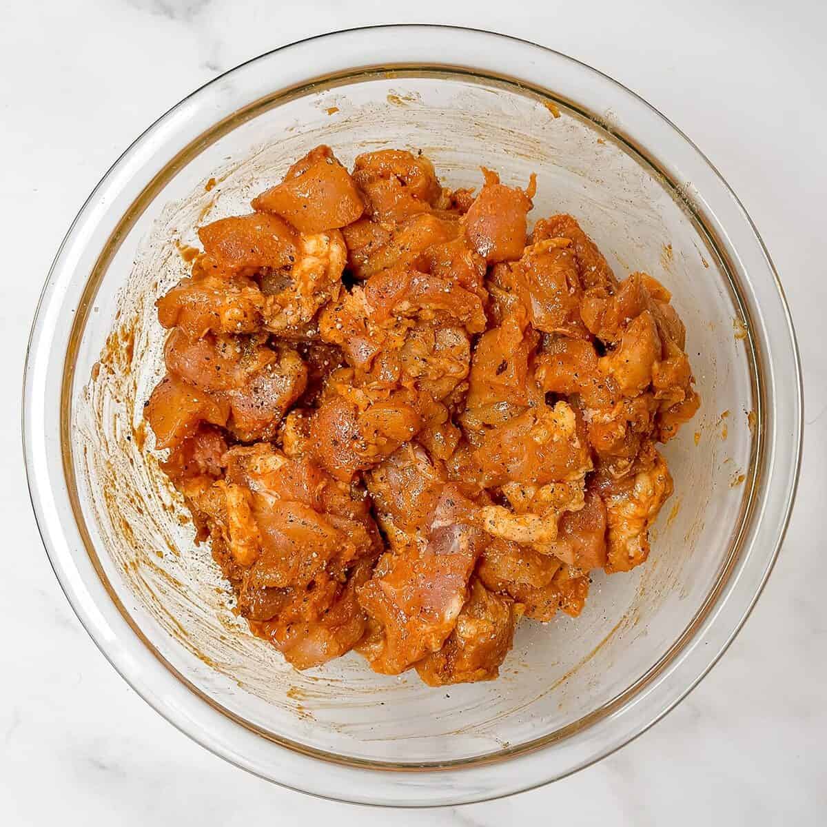 A glass bowl filled with raw, diced chicken breast marinated in spices for Chicken Shawarma on a white marble surface.