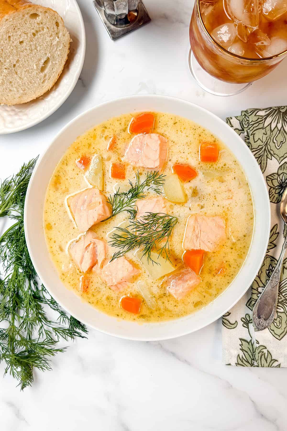 A bowl of creamy Finnish Salmon Soup with chunks of salmon, carrots, and potatoes, garnished with dill, sits on a white table next to bread, a drink, and a napkin with a spoon.
