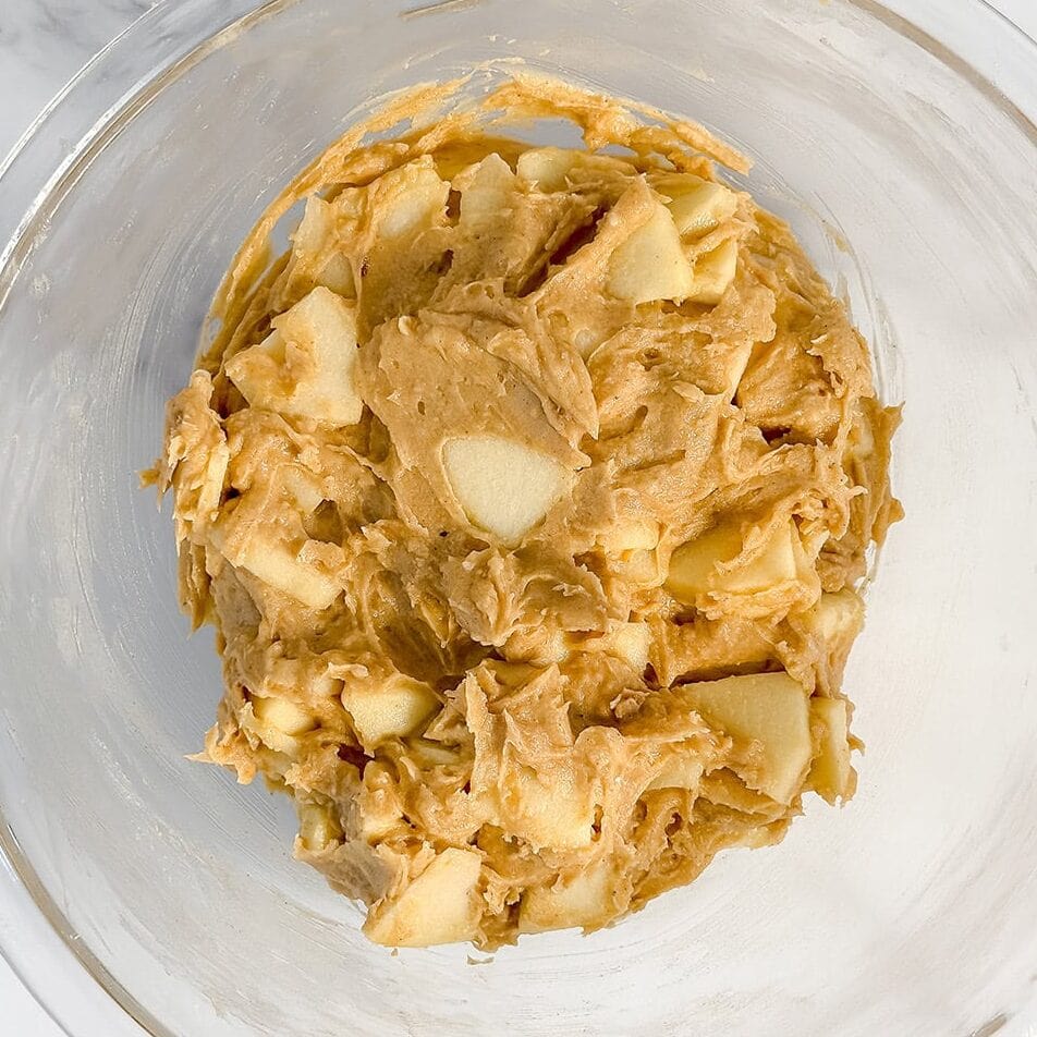 A clear glass bowl contains cookie dough with visible chunks of apple, ready to be baked into a delicious apple crumble cake, set on a white marble surface.