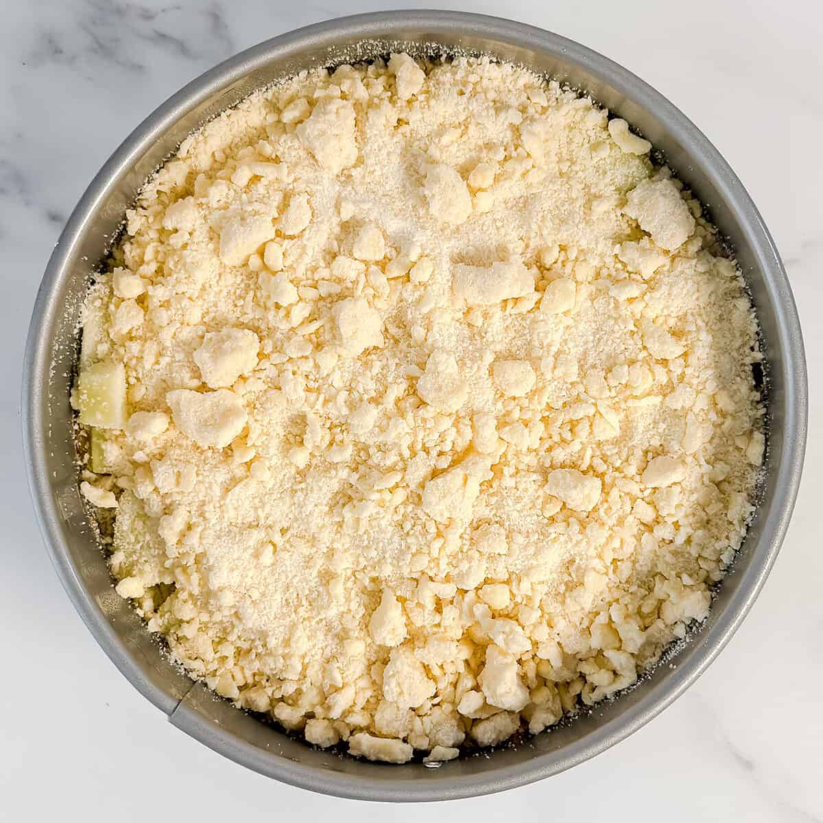 A round baking pan filled with an even layer of crumbly streusel topping sits atop a light marble surface, ready to become a delicious apple crumble cake.