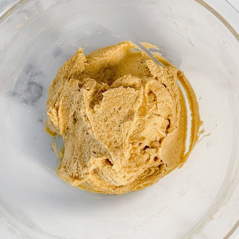 A clear glass bowl containing a portion of light brown apple crumble cake dough mixture on a white marble surface.