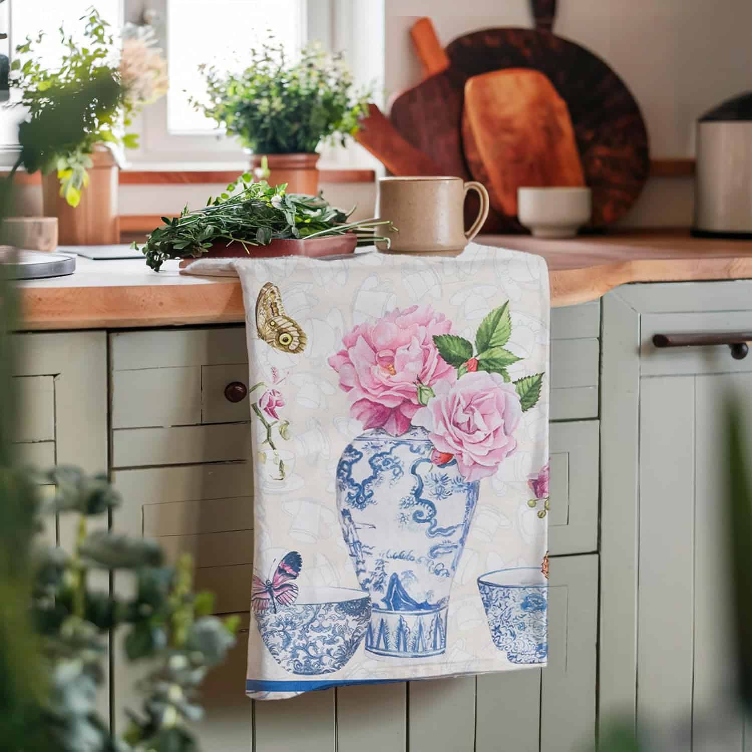 A floral tea towel with a blue and white vase design hangs over a light green kitchen cabinet-perfect for brightening up your kitchen on game day; a cup and herbs sit on the wooden countertop.