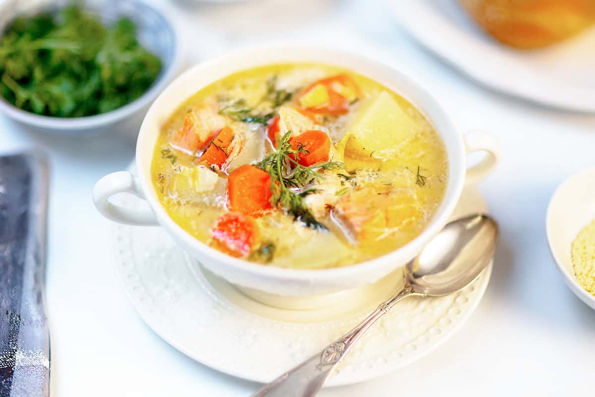 A bowl of Finnish Salmon Soup (Lohikeitto) with carrots, potatoes, herbs, and broth, served with a spoon on a white plate.