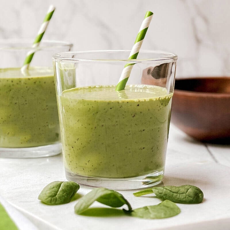 Two glasses of green smoothie with striped straws rest on a white surface, featuring fresh spinach leaves in front-perfect for enjoying your first greens-with a wooden bowl in the background.