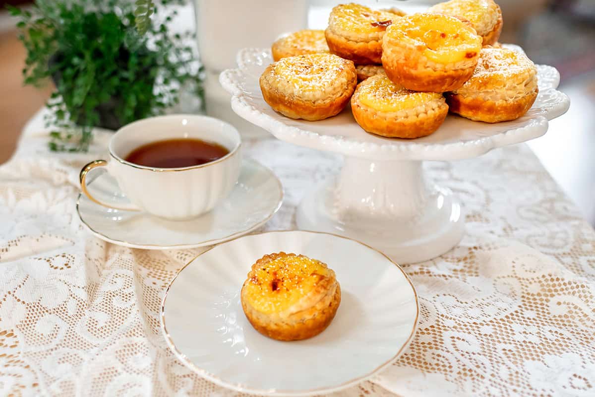 A plate of Irish Custard Tarts on a white cake stand, with one tart on a separate plate and a cup of tea on a lace tablecloth-a classic Irish dessert scene.